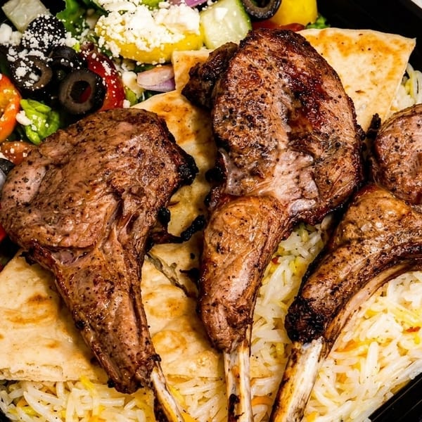 Four grilled lamb chops with rice, pita bread, and a fresh Greek salad in a black container.