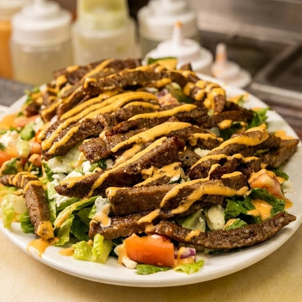 A large plate of steak salad with lettuce, tomatoes, and feta cheese, drizzled with sauce.