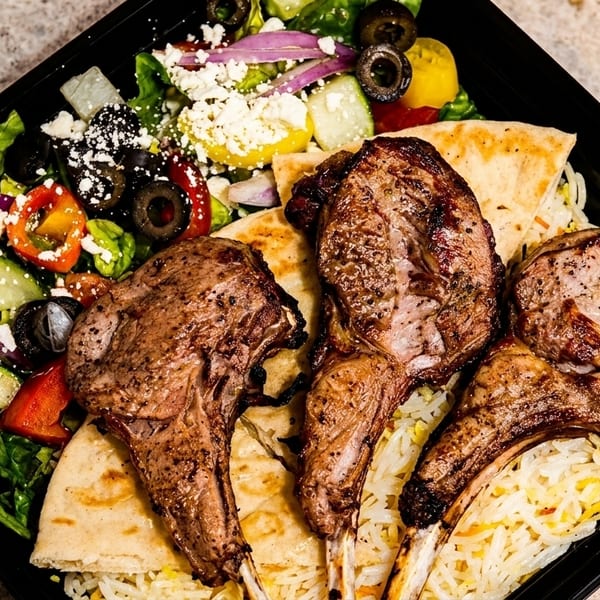 Grilled lamb chops, rice, pita bread, and a Greek salad with feta cheese in a black container.